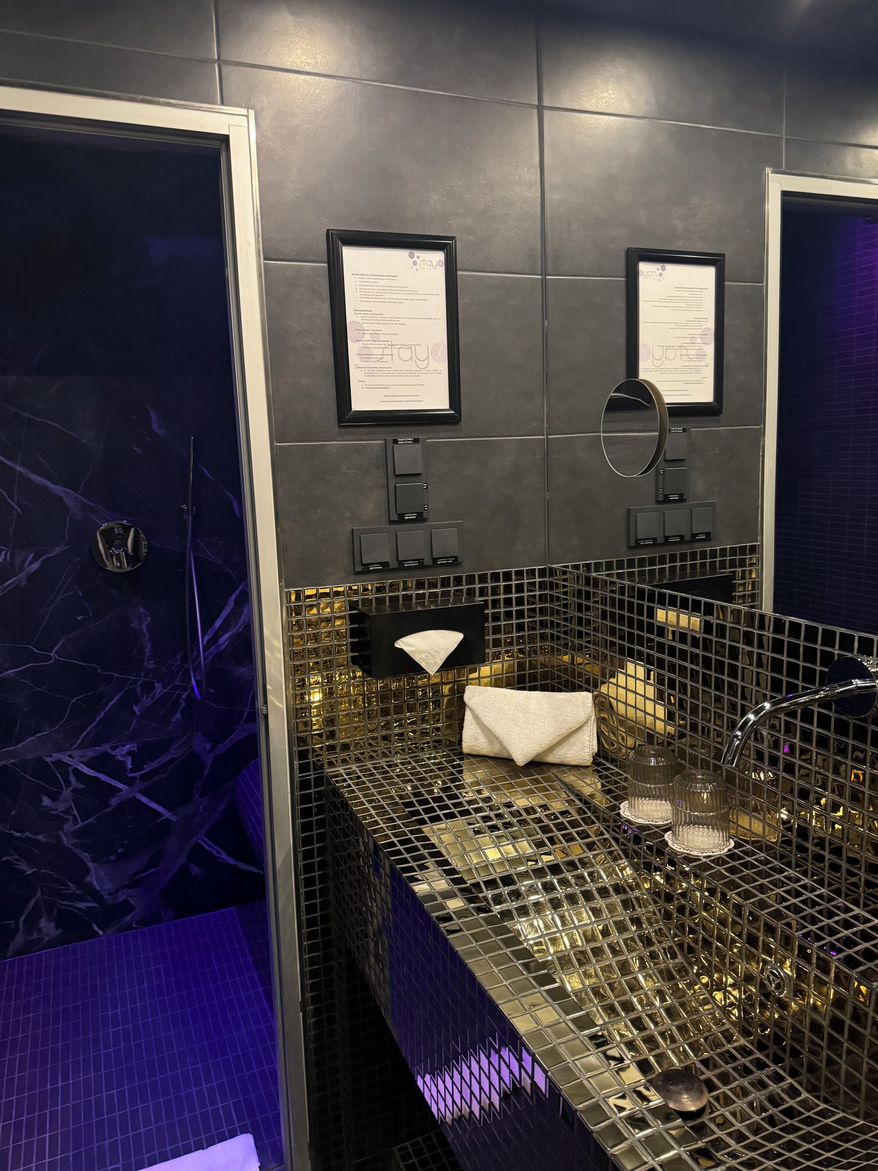 A modern bathroom with a marble wall, a metal sink with a grid pattern, and two framed certificates on the wall.