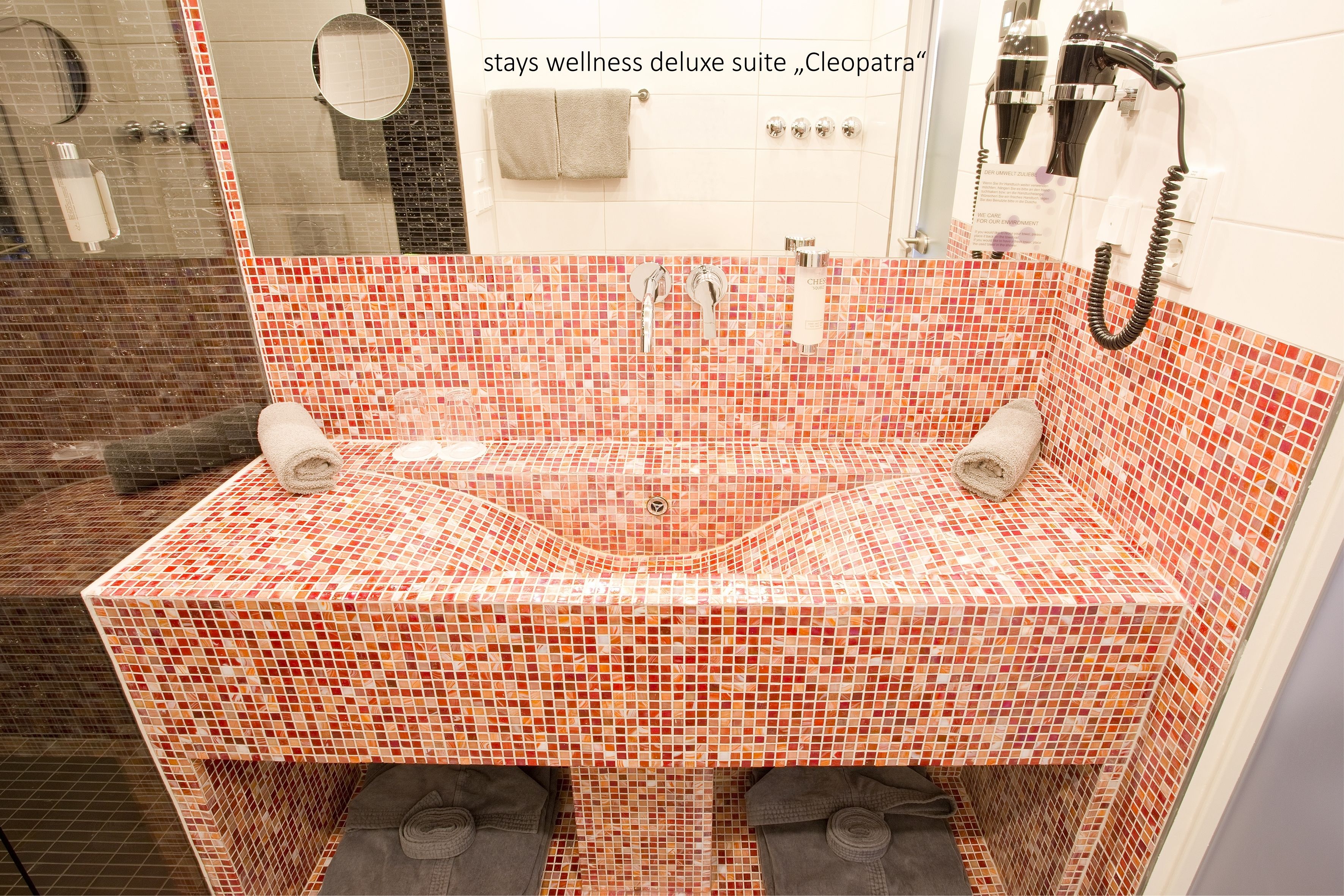 Ein Badezimmer mit einem Waschbecken aus Mosaikfliesen, zwei Handtuchhaltern, einer Duschkabine aus Glas und einem an der Wand befestigten Fön.
