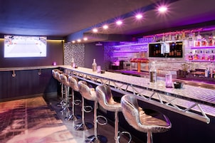 Zona de bar con una barra larga, varios taburetes y una pantalla mostrando un evento deportivo.