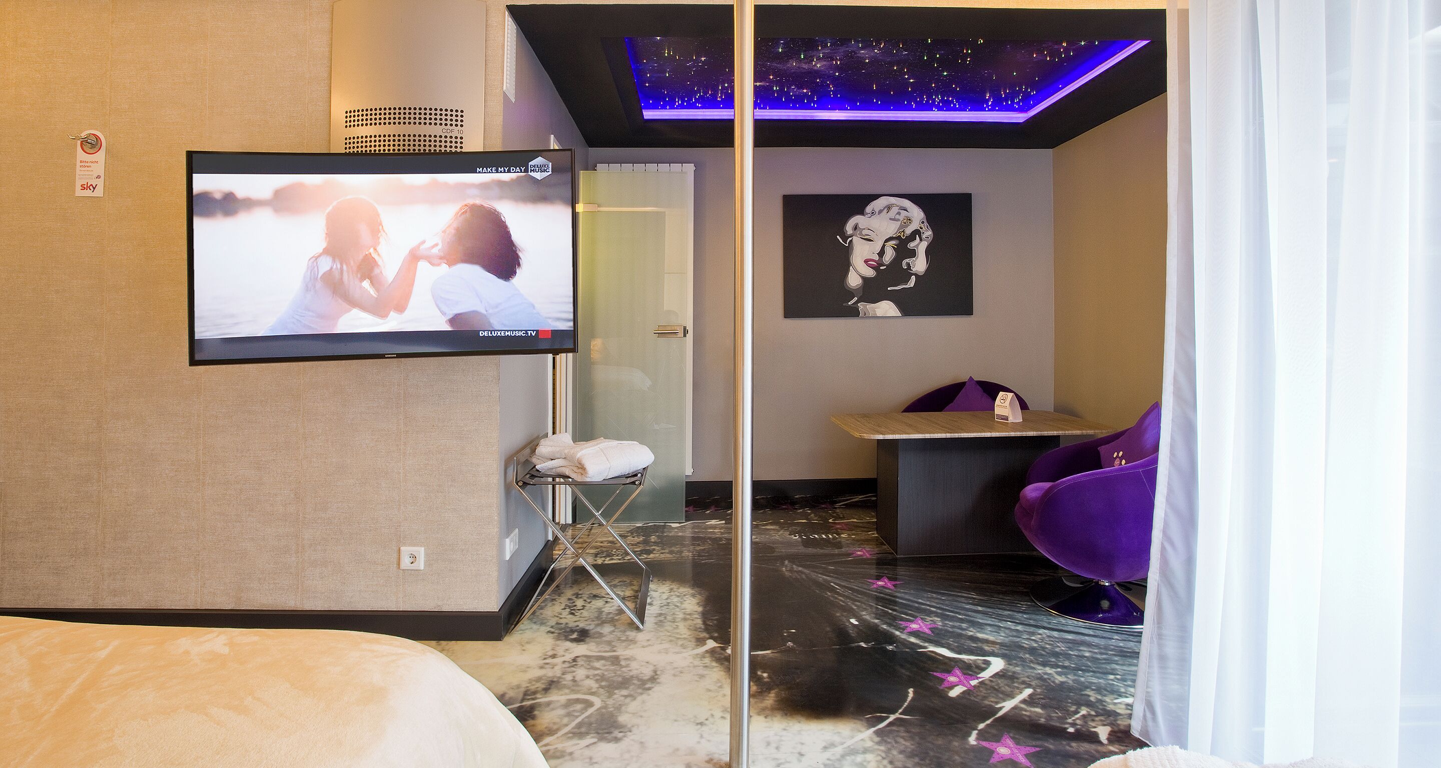 Une chambre d’hôtel équipée d’une télévision diffusant un film, d’un lit, d’un coin salon avec une table et des chaises, et d’un mur orné d’un tableau encadré.