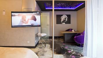 Une chambre d’hôtel équipée d’une télévision diffusant un film, d’un lit, d’un coin salon avec une table et des chaises, et d’un mur orné d’un tableau encadré.