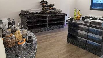 A breakfast bar with various food items and condiments, a coffee station, and a fruit basket.