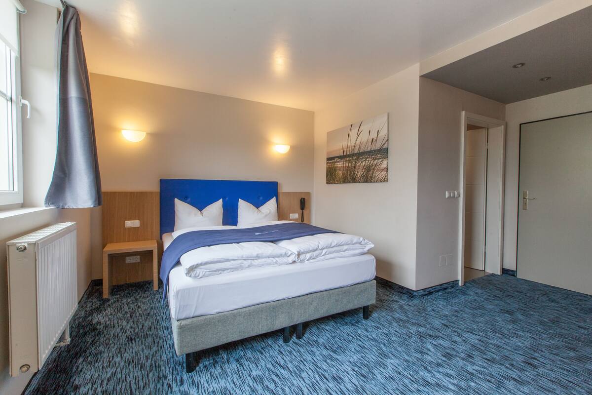 A hotel room with a bed, bedside tables, a radiator, a blue carpet, and a wall-mounted artwork.