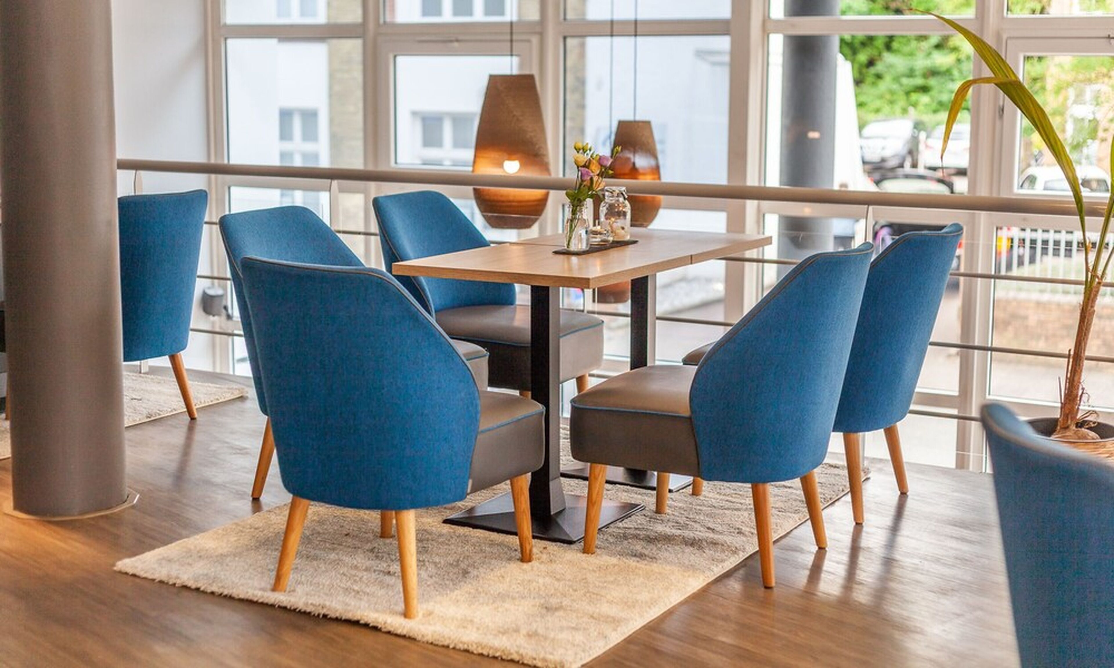 A modern dining area with blue upholstered chairs, a wooden table, and a view through large windows.