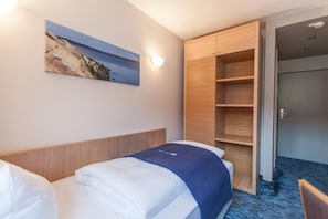 A hotel room with a bed, a wardrobe, a painting of a coastal scene, and a carpeted floor.