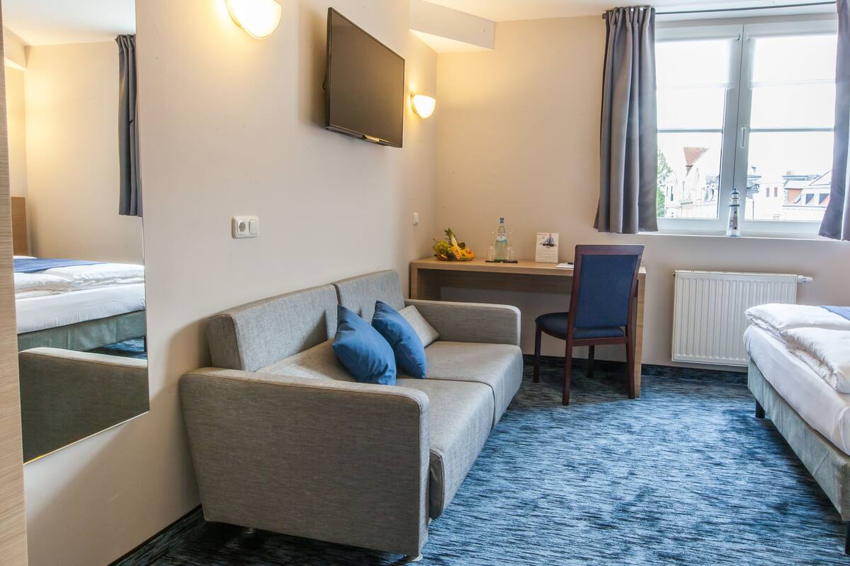A hotel room with a sofa, a desk, a chair, a bed, a TV, and a window with curtains.