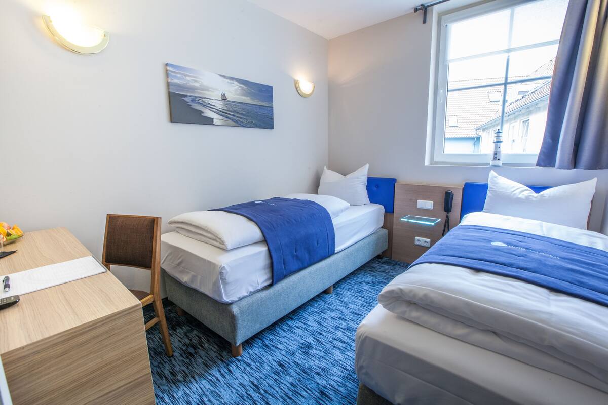 A hotel room with a bed, a desk, a chair, a bedside table, a lamp, a painting, and a window with curtains.