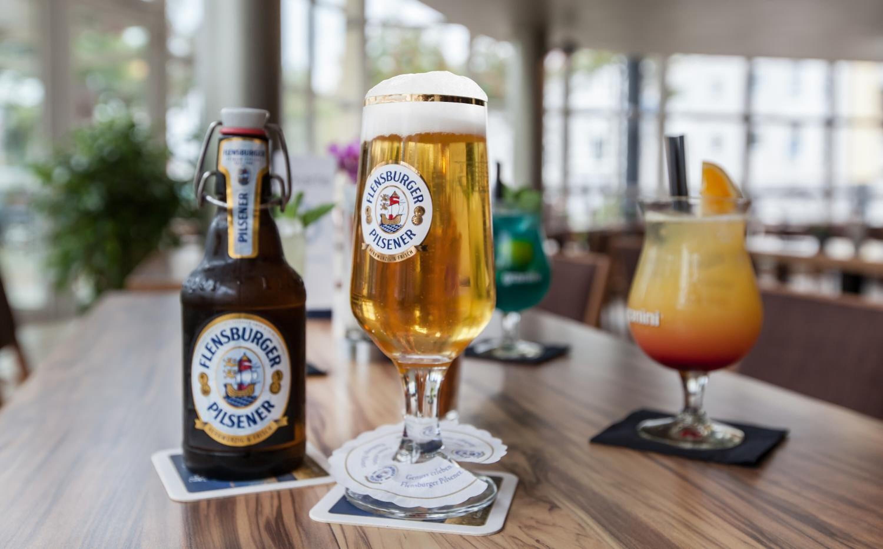A bottle of pilsner beer, a glass of pilsner beer, and a cocktail on a wooden table.