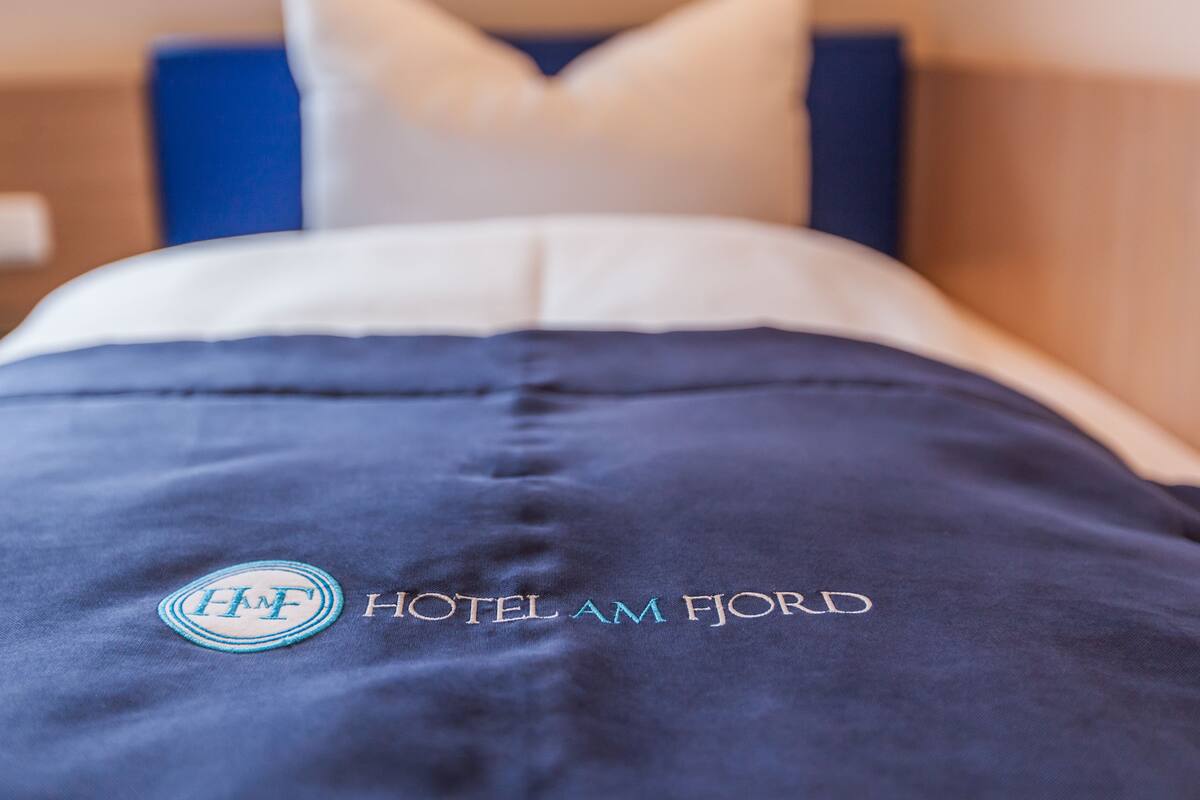 A hotel bed with a blue bedspread and pillows, and the logo and name of the hotel, HOTEL AM FJORD, embroidered on the bedspread.