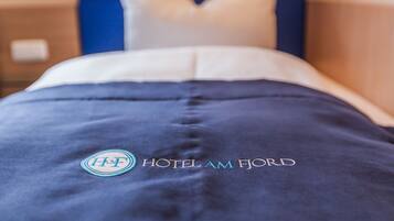 A hotel bed with a blue bedspread and pillows, and the logo and name of the hotel, HOTEL AM FJORD, embroidered on the bedspread.