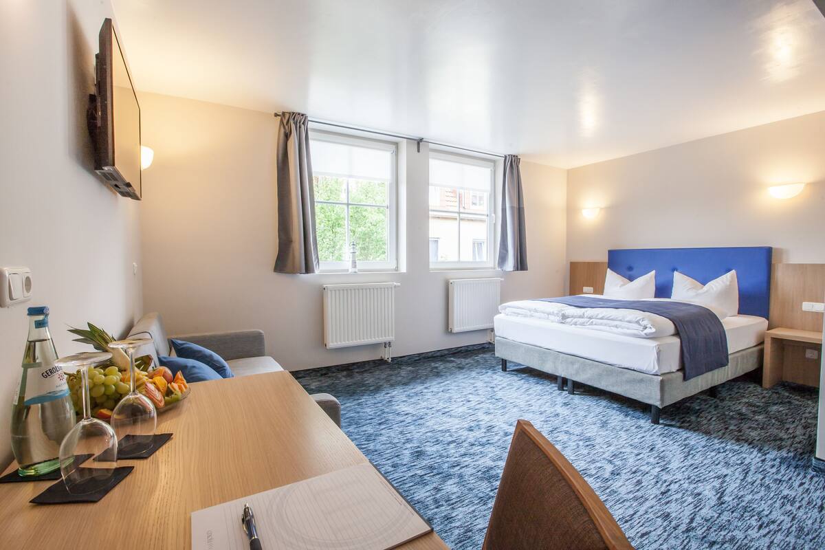 A hotel room with a bed, bedside tables, a desk with a chair, a dining table with a chair, a TV mounted on the wall, and windows with curtains.