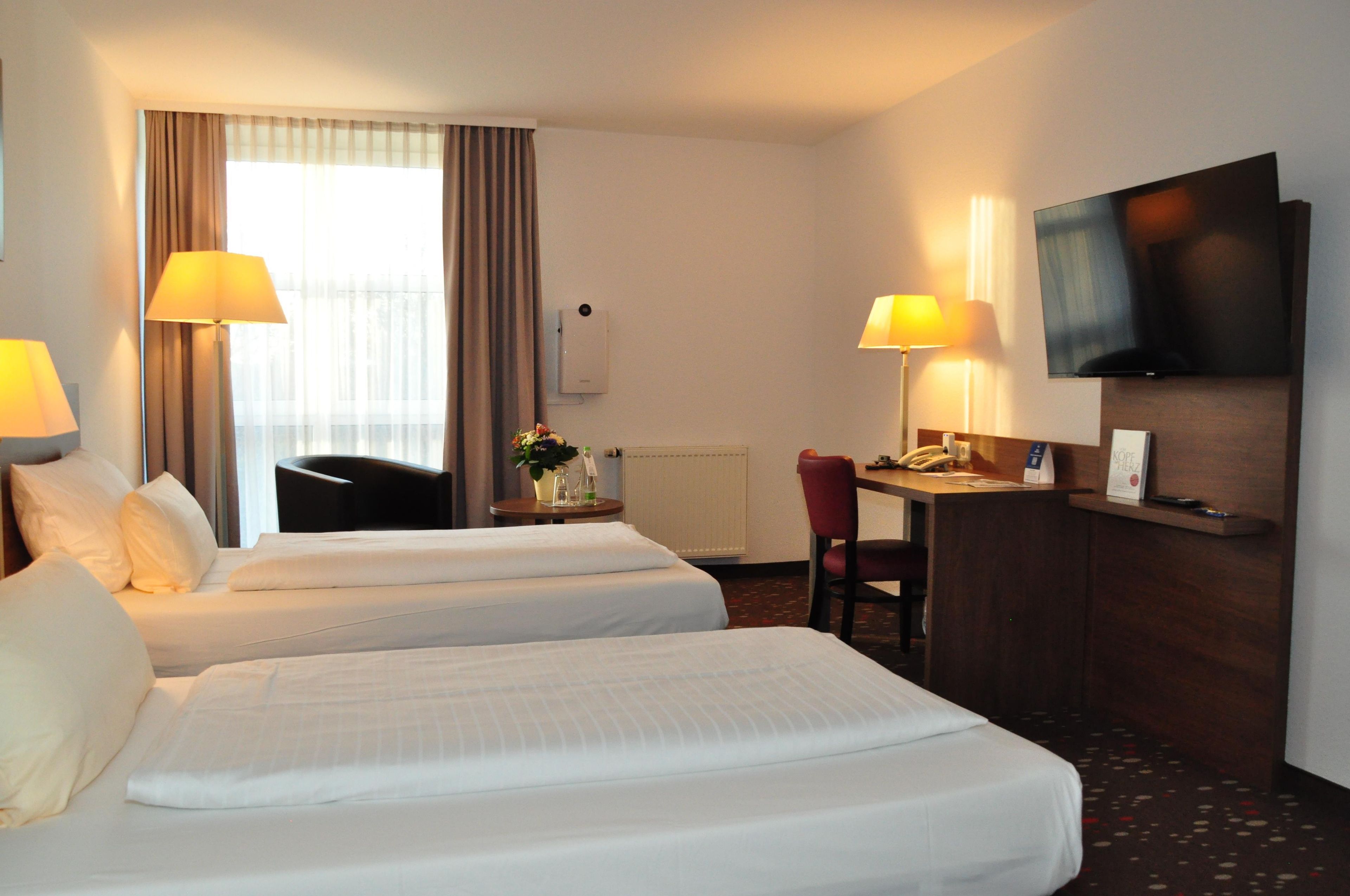 Une chambre d’hôtel avec deux lits, un bureau, une chaise, une télévision et une fenêtre avec des rideaux.