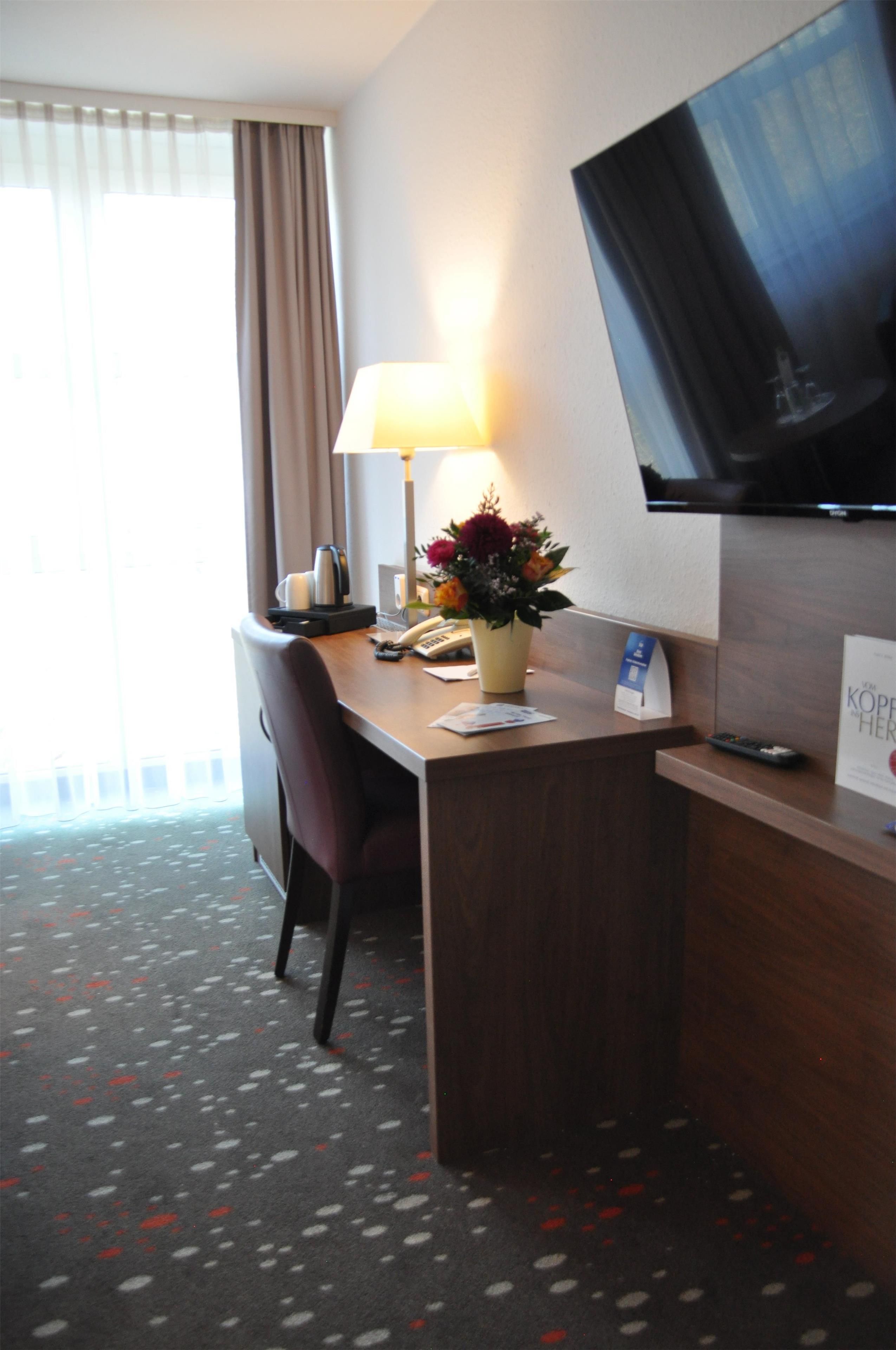 Ein Hotelzimmer mit Schreibtisch, Stuhl, Lampe, Blumenvase, Wandfernseher und einem Vorhangfenster.