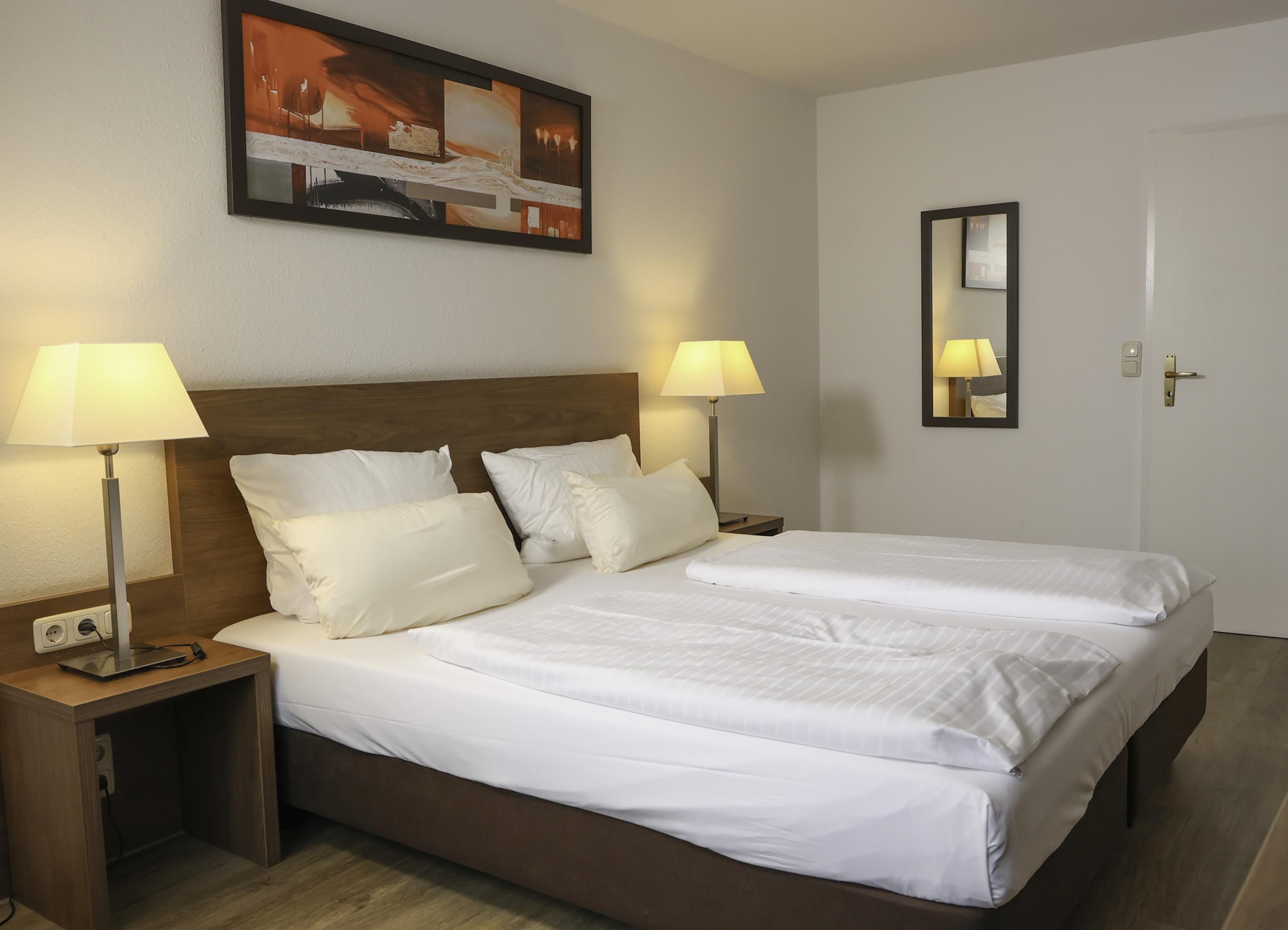 Ein Hotelzimmer mit Doppelbett, zwei Nachttischen inklusive Lampen, einem Bild an der Wand und einer Tür mit Spiegel.