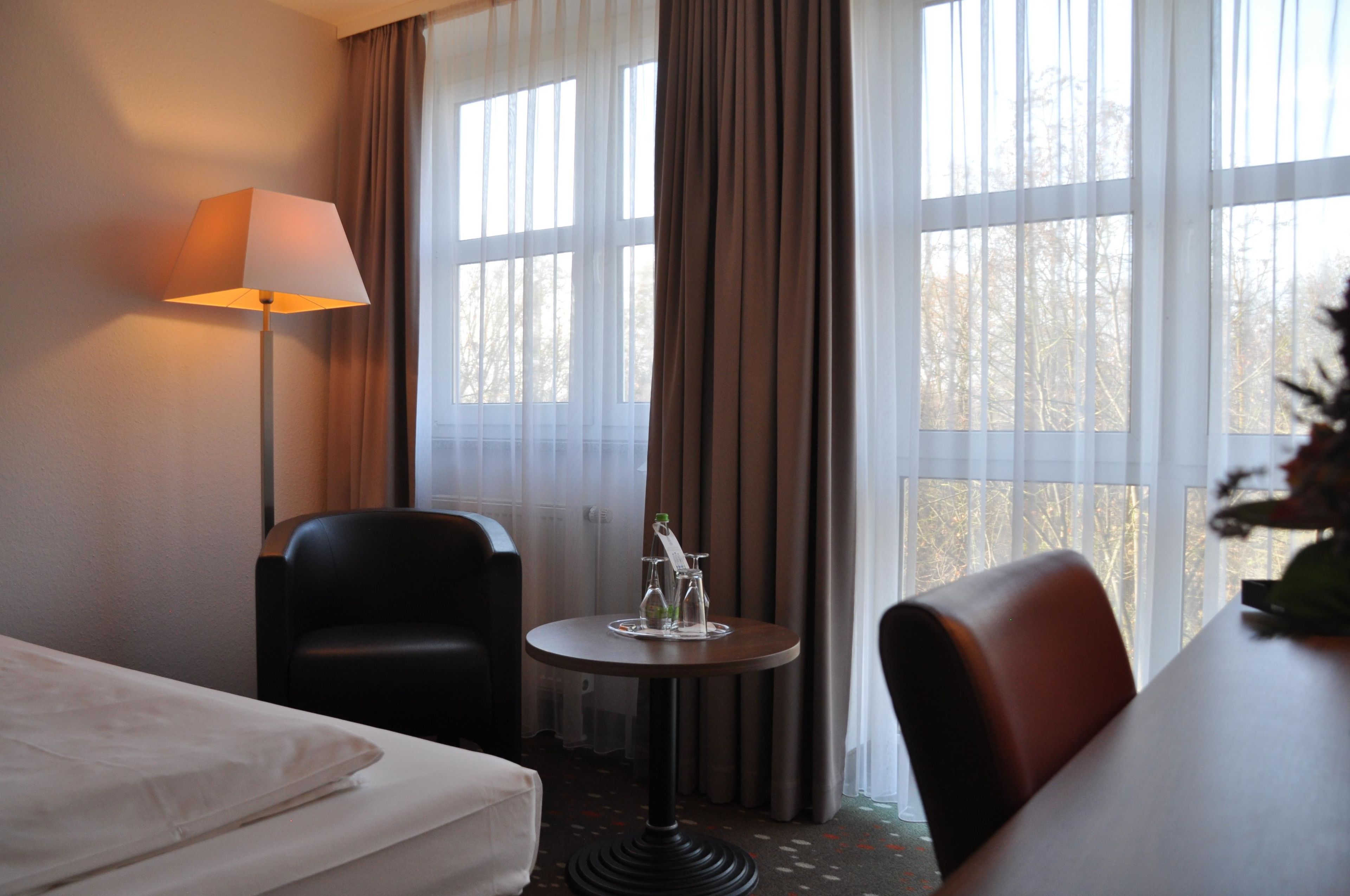 Habitación de hotel con cama, escritorio, dos sillas, lámpara y ventana con cortinas.