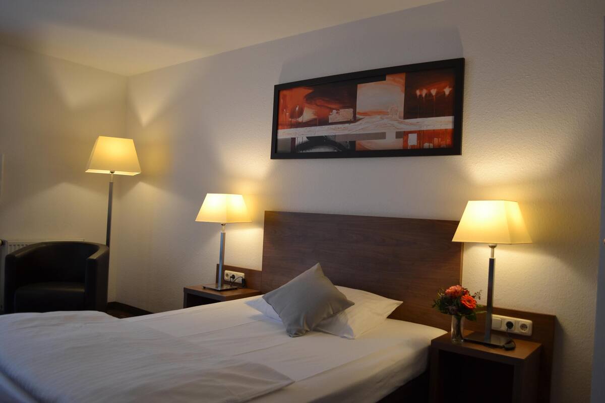 A hotel room with a large bed, two bedside lamps, a nightstand with a vase of flowers, and a framed picture on the wall.