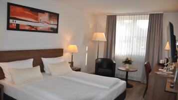 Habitación de hotel con una cama grande, dos lámparas de noche, un escritorio con una silla, una mesita con lámpara y un cuadro en la pared.