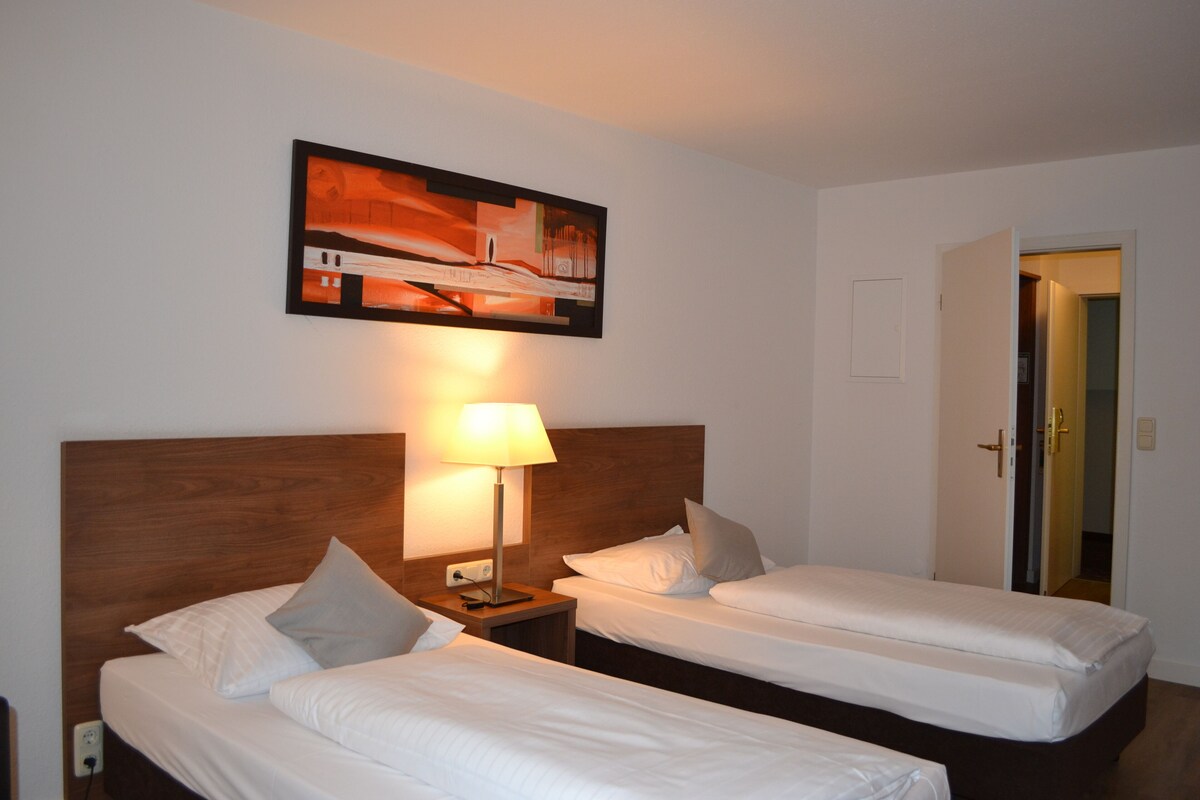 A hotel room with two single beds, a wooden headboard, a nightstand with a lamp, and a painting on the wall.