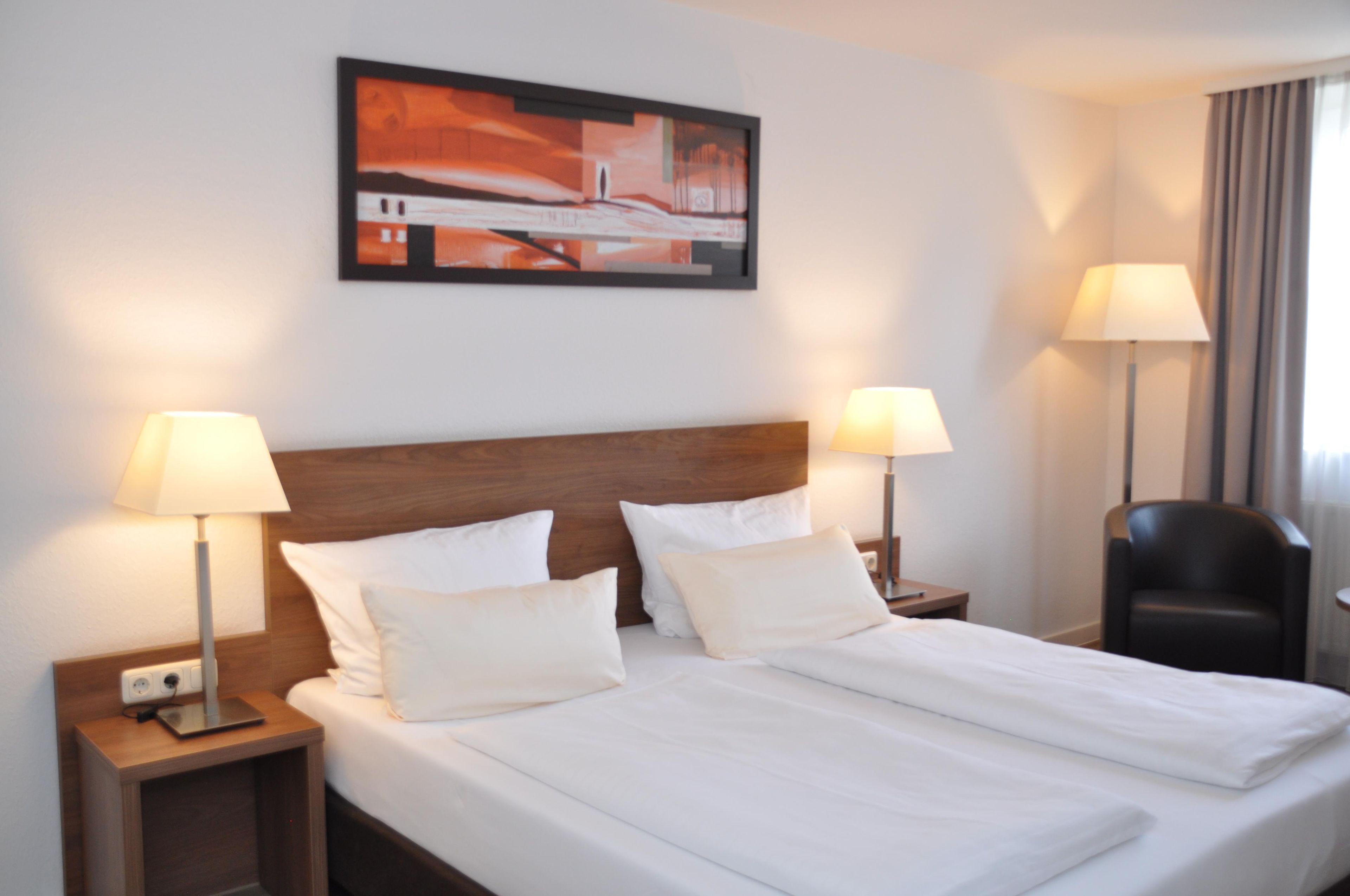 A hotel room with a large bed, two bedside lamps, a headboard, a nightstand, a chair, and a painting on the wall.