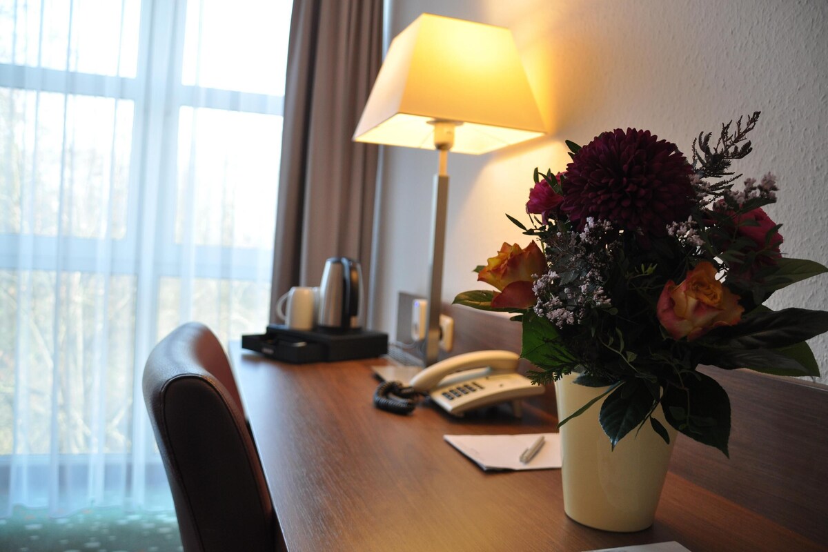 Habitación de hotel con escritorio de madera, lámpara, teléfono y un florero con flores.