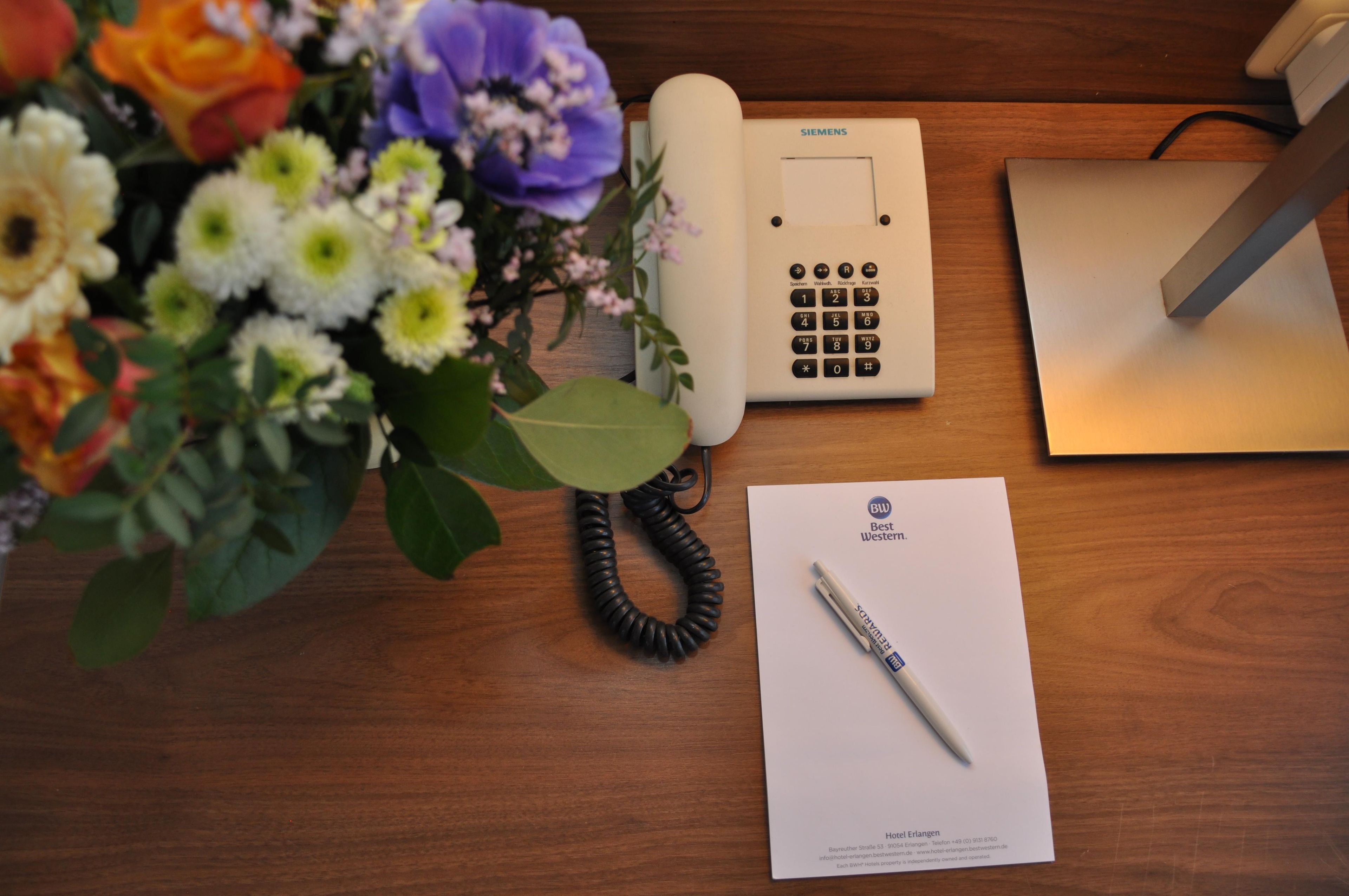 A wooden desk with a Siemens telephone, a pen, a paper with hotel information, and a bouquet of flowers.
