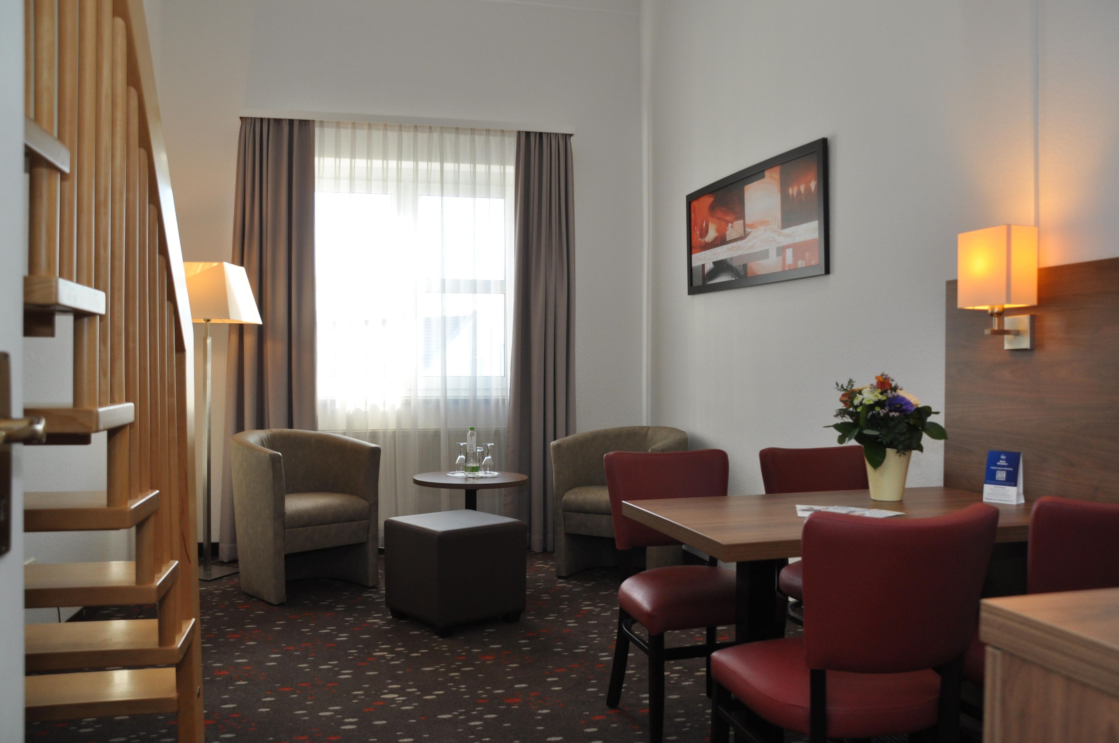 Habitación de hotel con una escalera, dos sillones, una mesita con un florero, una mesa de comedor con sillas, un cuadro en la pared y una ventana con cortinas.