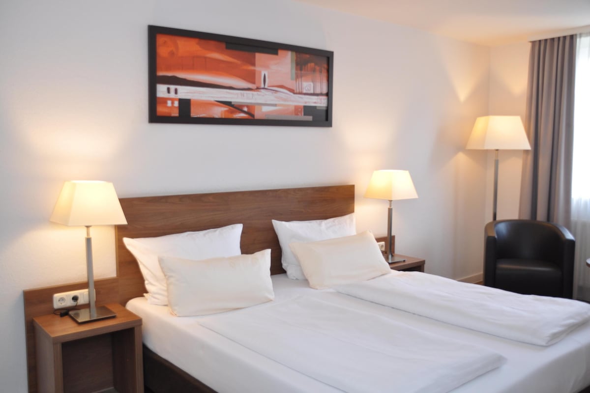 A hotel room with a large bed, two bedside lamps, a headboard, a nightstand, a chair, and a painting on the wall.
