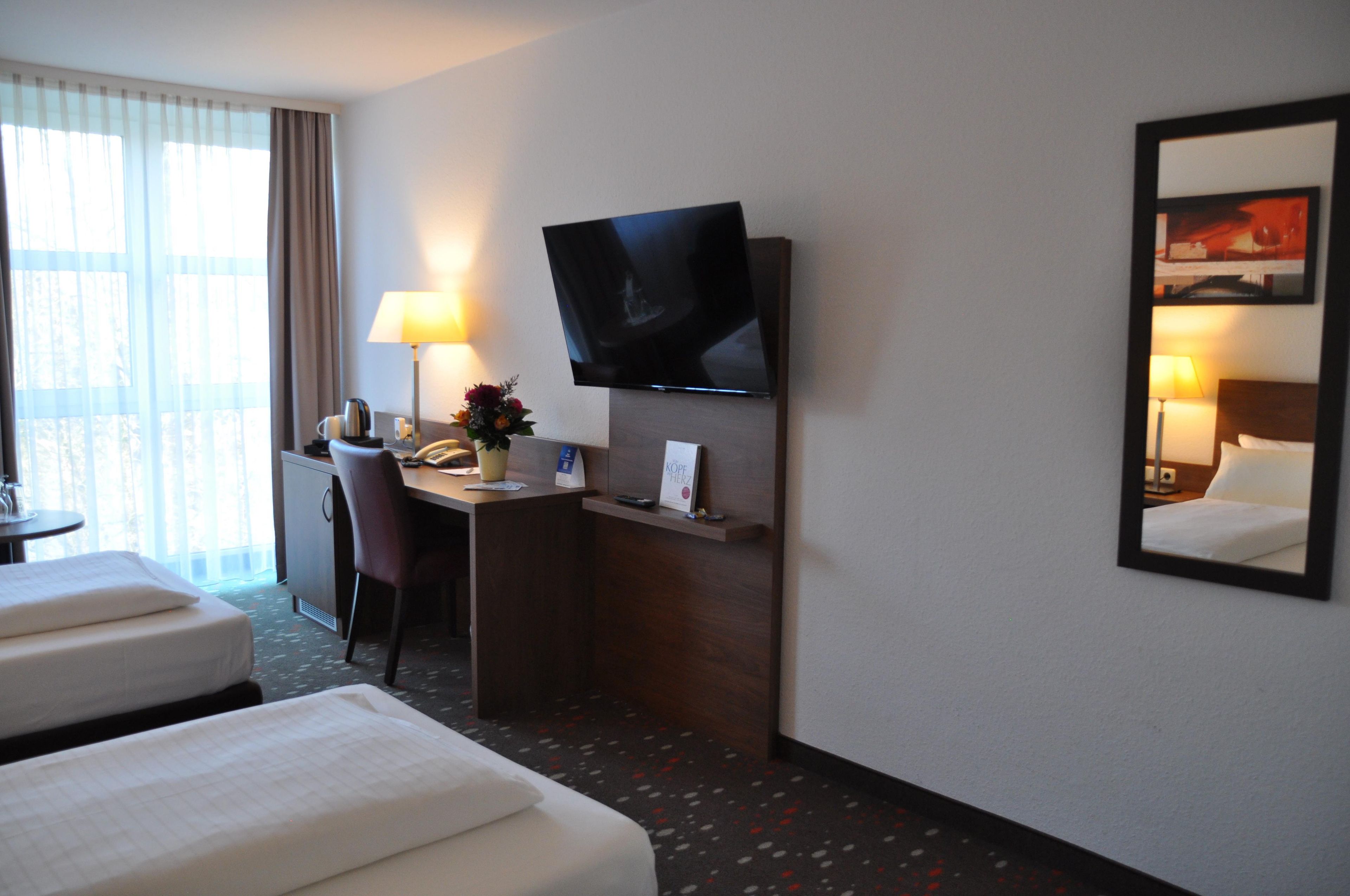 Una habitación de hotel con una cama, un escritorio, una silla, un televisor montado en la pared y un ventanal con cortinas.