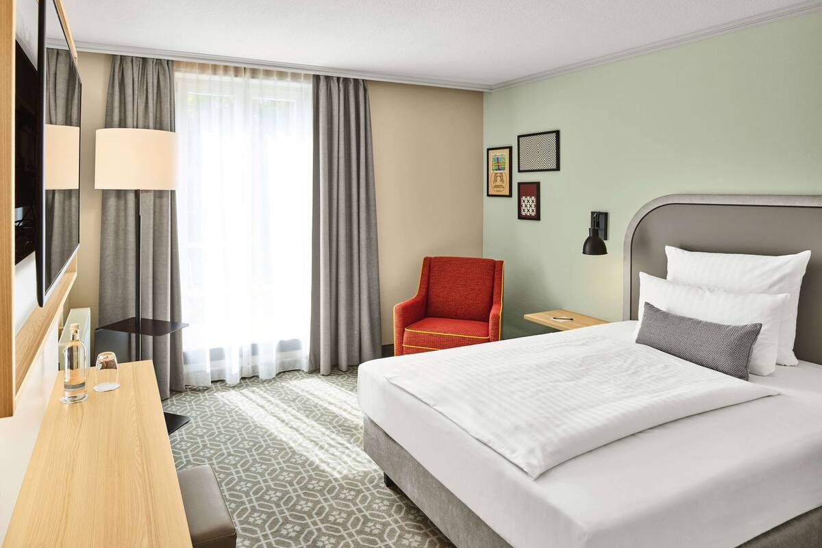 Ein Hotelzimmer mit Bett, Schreibtisch, Stuhl, Fernseher und einem Fenster mit Vorhängen.