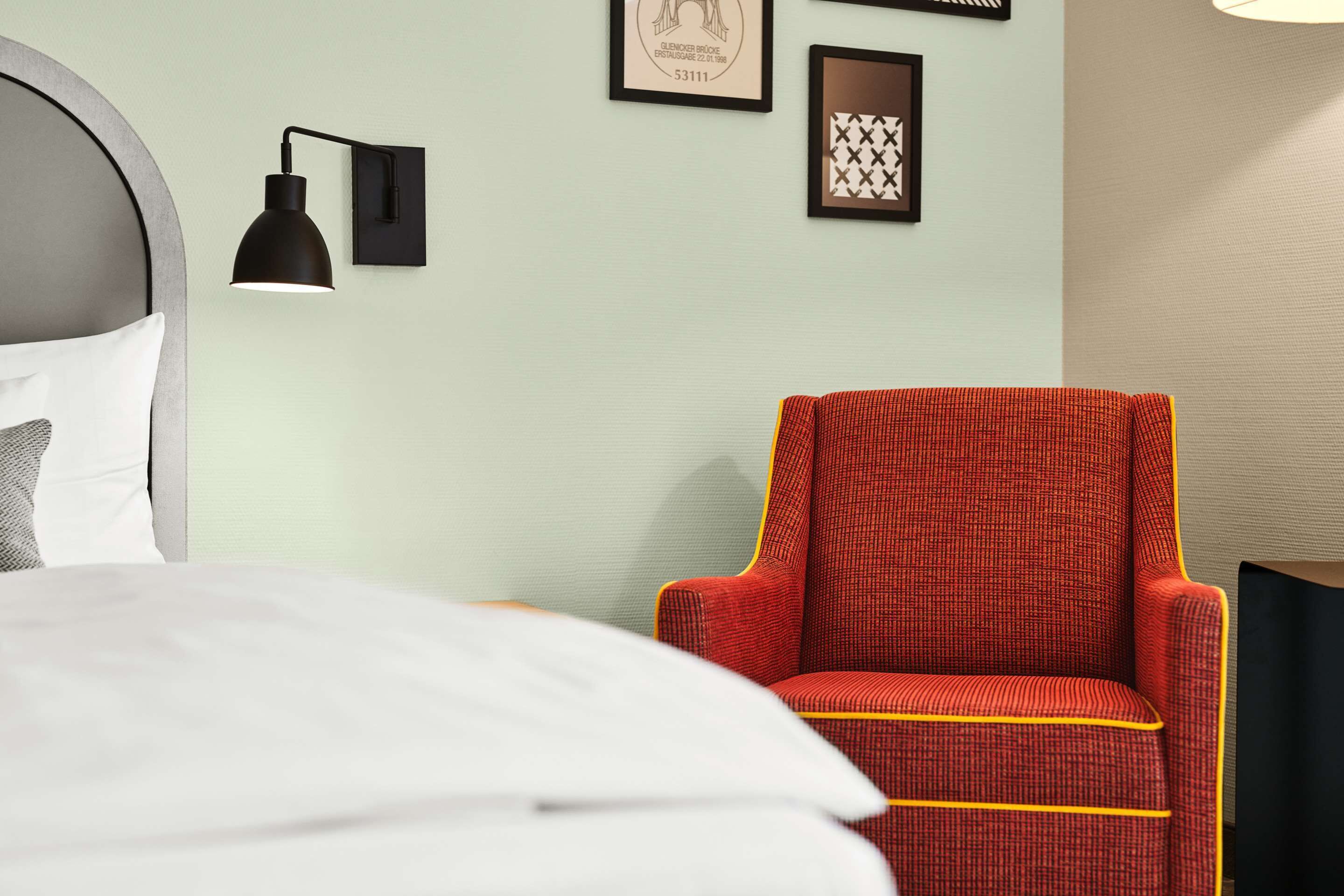 A hotel room with a bed, a red armchair, a black wall lamp, and two framed pictures on the wall.