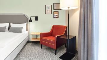 A hotel room with a bed, a red armchair, a small table, a floor lamp, and patterned carpet.