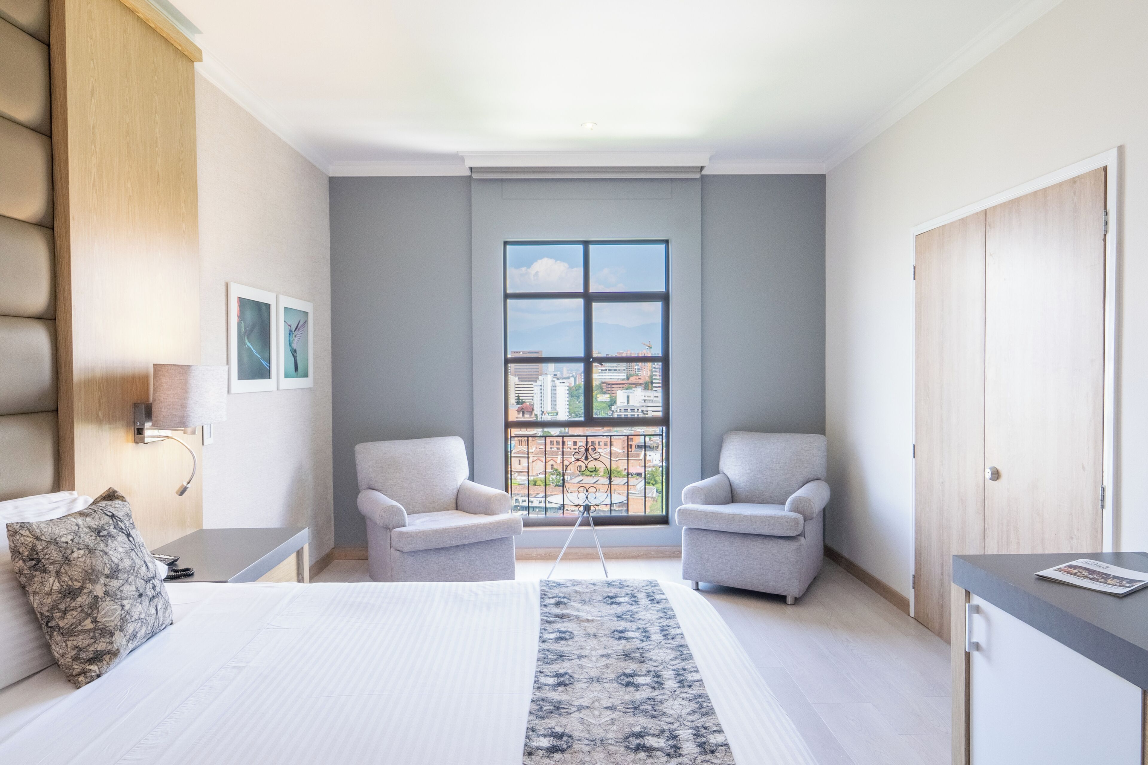 Habitación de hotel moderna con una cama grande, dos sillones, un escritorio, un ventanal con vistas a la ciudad y un balcón.