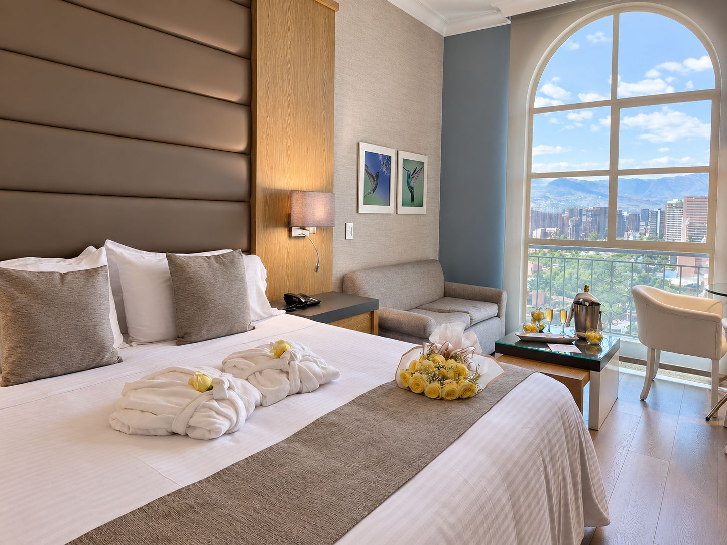 A hotel room with a large bed, a sofa, a desk with a chair, and a view of the city through a large window.
