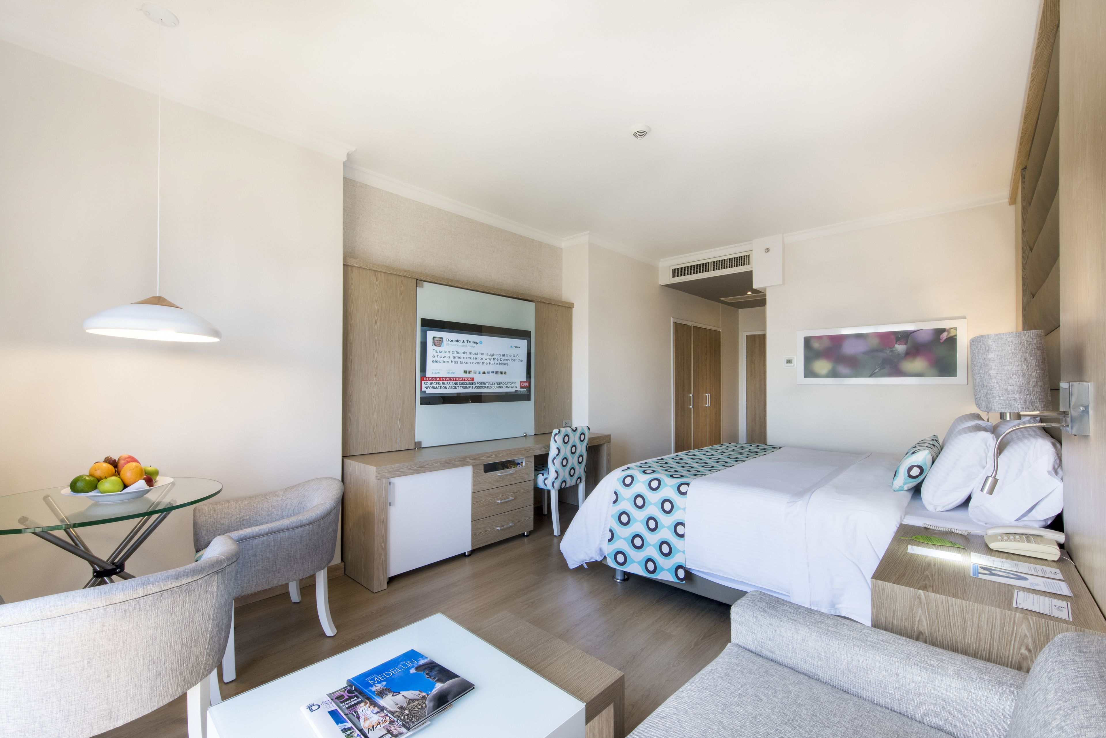 Habitación de hotel con cama, escritorio con computadora, silla, mesita de vidrio con fruta y televisor en la pared.