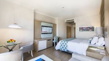 Habitación de hotel con cama, escritorio con computadora, silla, mesita de vidrio con fruta y televisor en la pared.