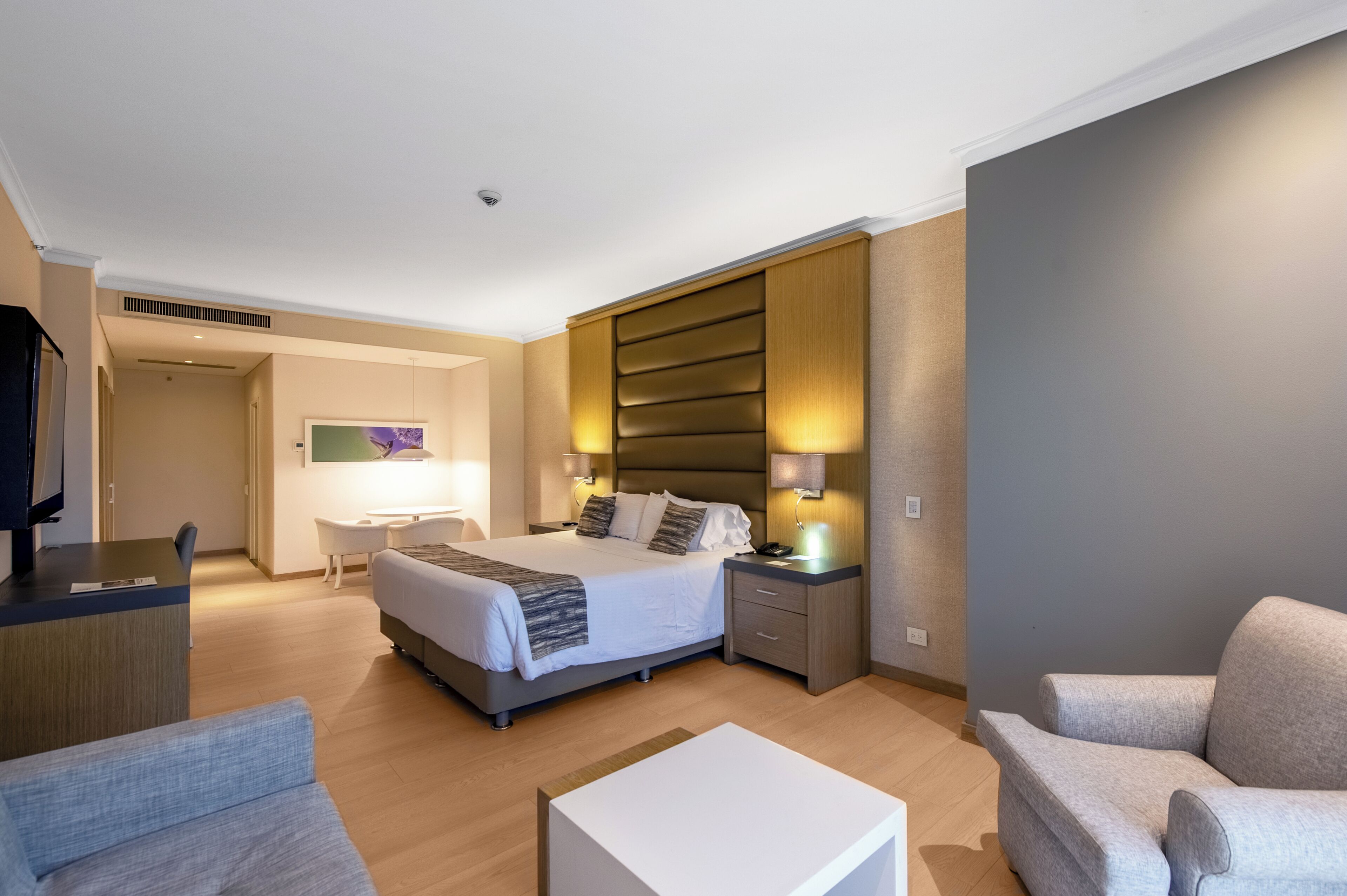 Ein modernes Hotelzimmer mit Bett, Nachttischen, Schreibtisch, Sessel und Fernseher.