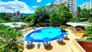 A rooftop pool with lounge chairs and umbrellas, surrounded by greenery and tall buildings.