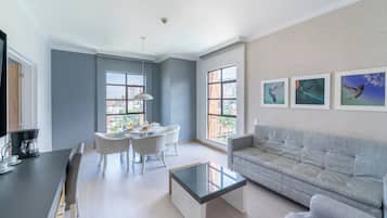 A modern living room with a sofa, a glass coffee table, a dining area with chairs, and large windows offering a city view.