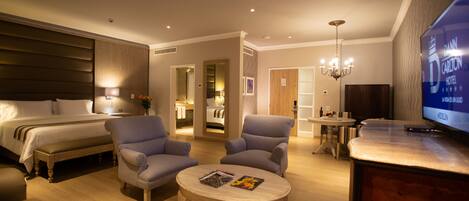 A modern hotel room with a large bed, two armchairs, a round coffee table, a TV, and a dining area with a table and chairs.