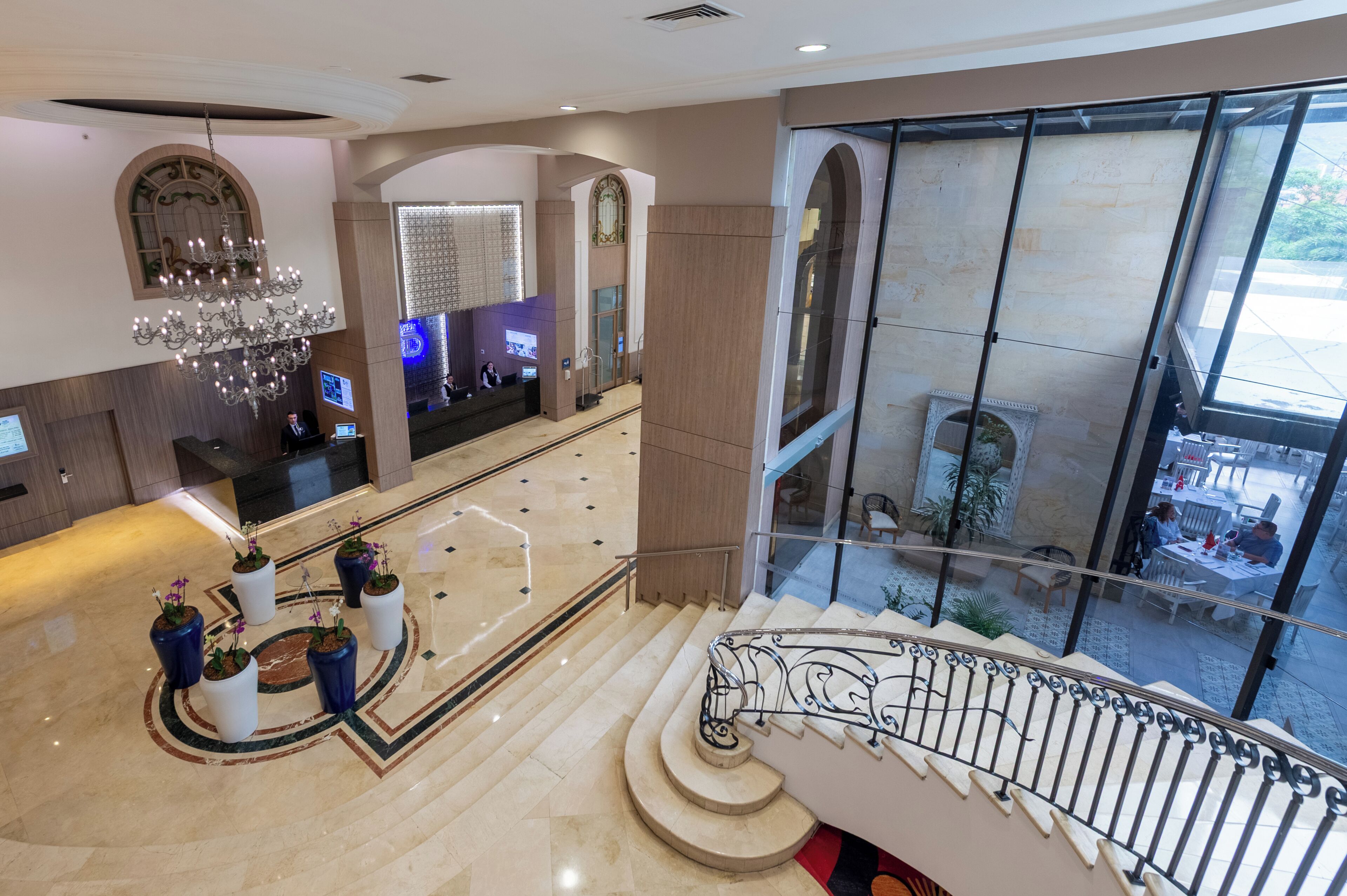 A hotel lobby with a grand staircase, a reception desk, and a view of the outdoor seating area.