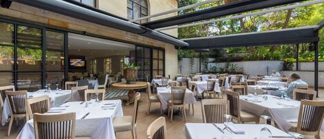An outdoor dining area with tables and chairs, large windows, and a view of the surrounding greenery.