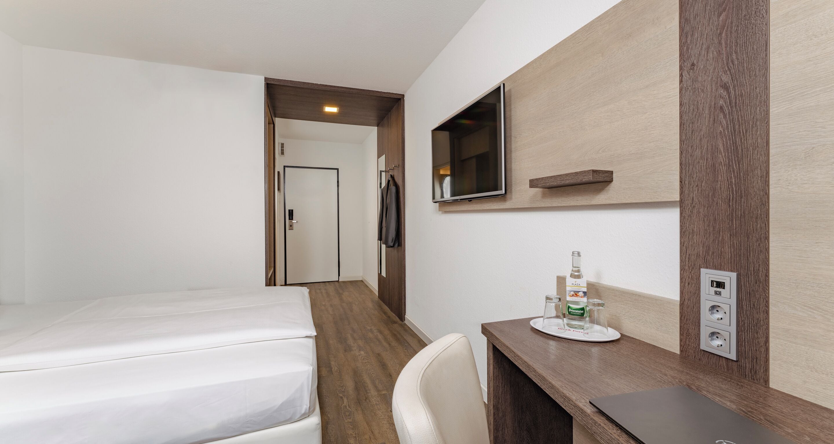 Ein Hotelzimmer mit einem Bett, einem Schreibtisch mit einer Wasserflasche, Gläsern und einem an der Wand befestigten Fernseher.