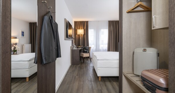 A hotel room with two beds, a desk with a chair, a minibar, a hanging coat, and a suitcase. - PLAZA INN Karlsruhe Nord (Karlsruhe)