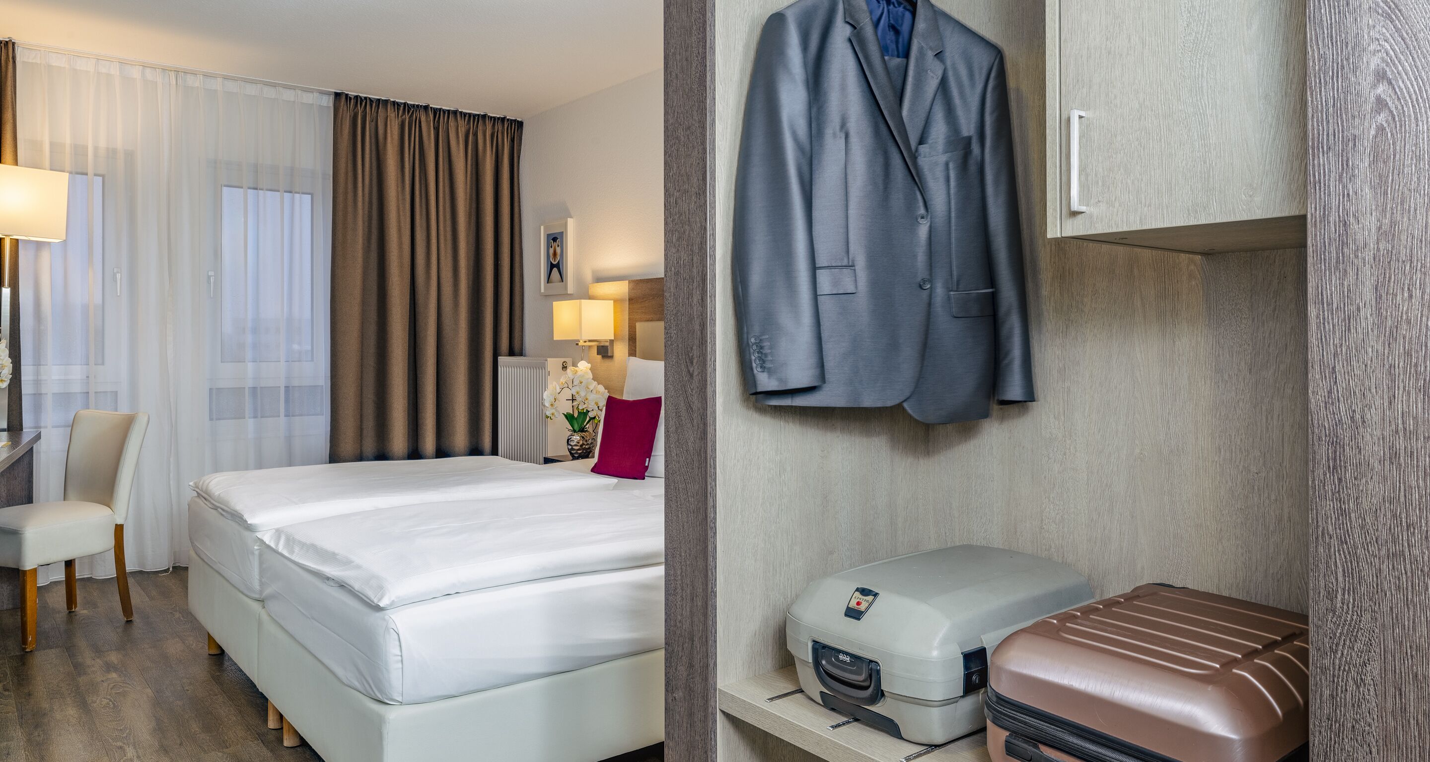 Ein Hotelzimmer mit Bett, Schreibtisch, Stuhl, Kleiderhaken und Koffer.