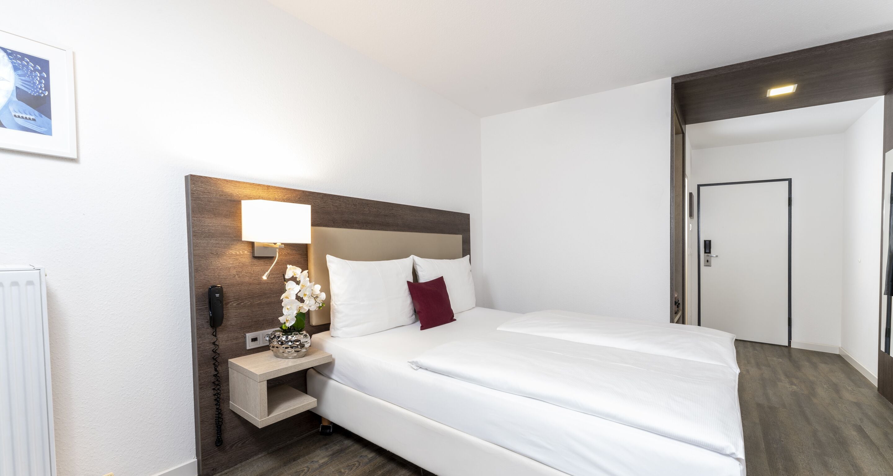 Ein Hotelzimmer mit einem großen Bett, einem Nachttisch mit Lampe, einer Vase mit Blumen und einer Tür.