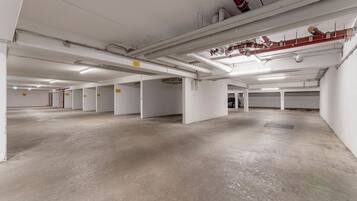 An empty parking garage with concrete floors and white walls, featuring multiple parking spaces and visible ceiling pipes.