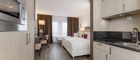 A hotel room with a kitchenette, a bed, a desk, and a chair.