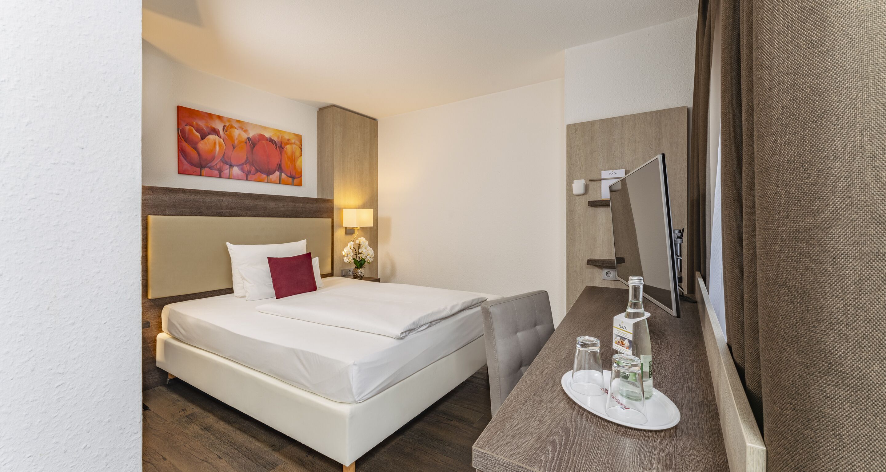 Ein Hotelzimmer mit Bett, Nachttisch mit Flasche und Glas, Spiegel und einem Gemälde an der Wand.
