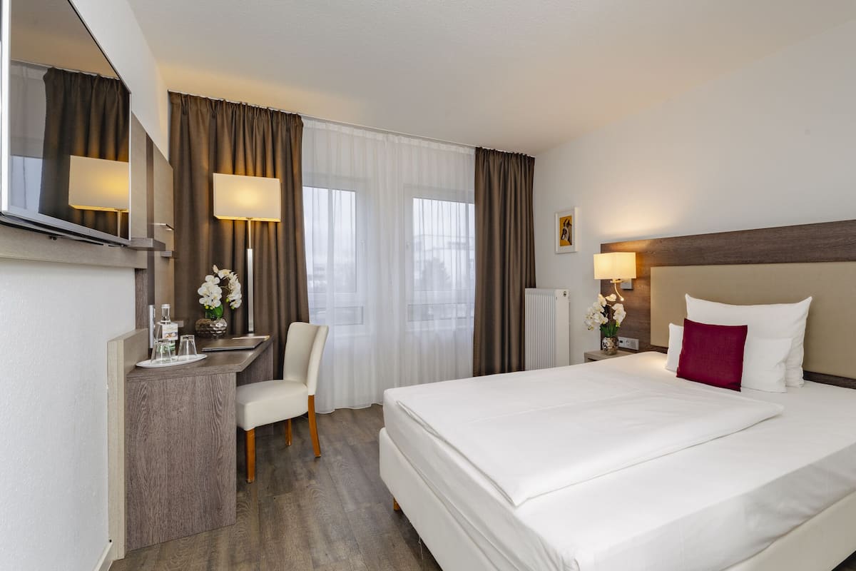 Habitación de hotel con una cama grande, un escritorio con silla, un espejo y una ventana con cortinas.