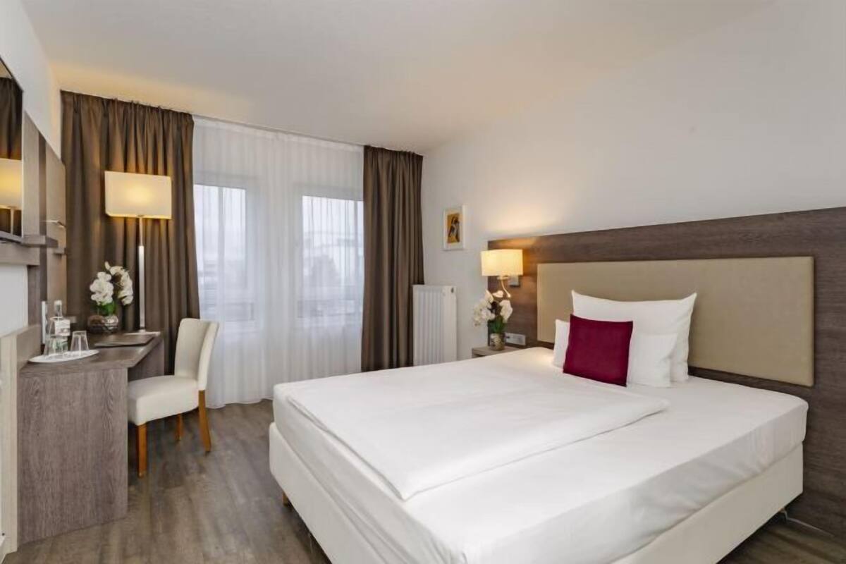 Habitación de hotel con una cama grande, un escritorio con silla, una lámpara y un ventanal con cortinas.